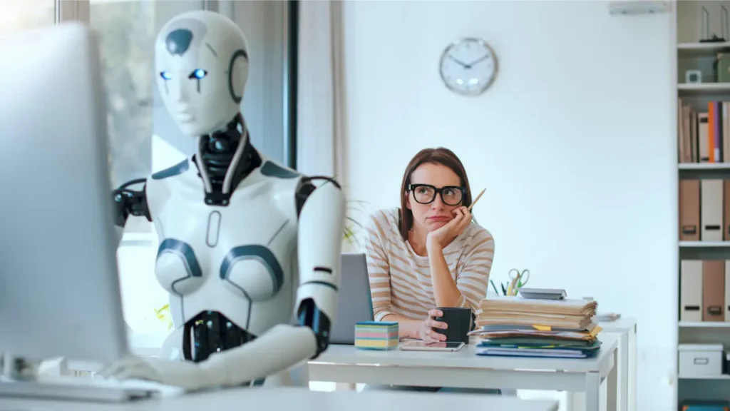 Humanoid AI robot working at a computer while an office employee observes, illustrating how artificial intelligence is changing the workplace and job market.